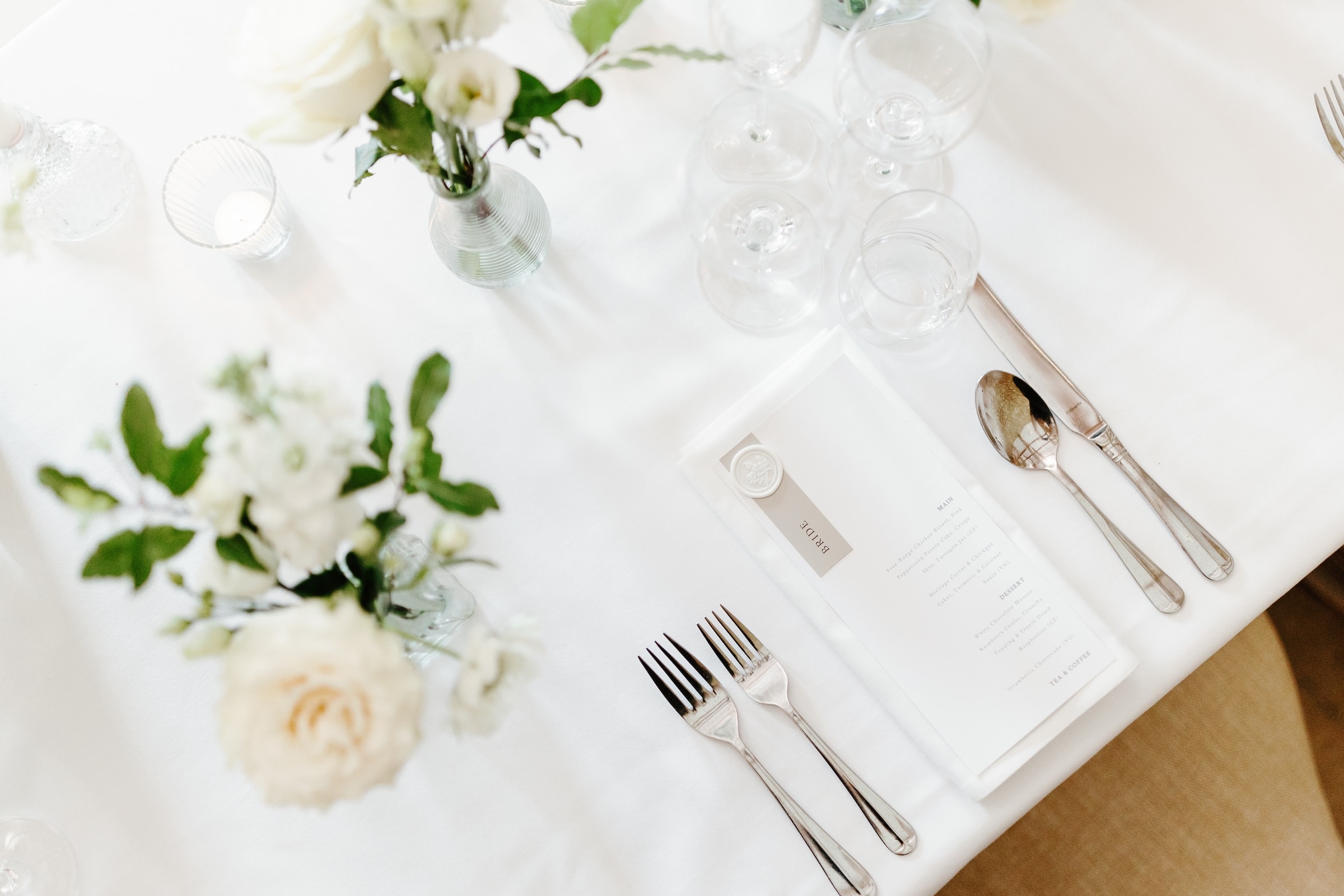 A formal table setting with a menu, cutlery, wine glasses, and white flower arrangements on a white tablecloth.