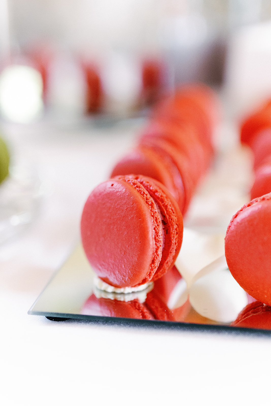 A row of red macarons is arranged on a reflective surface with blurred backgrounds.