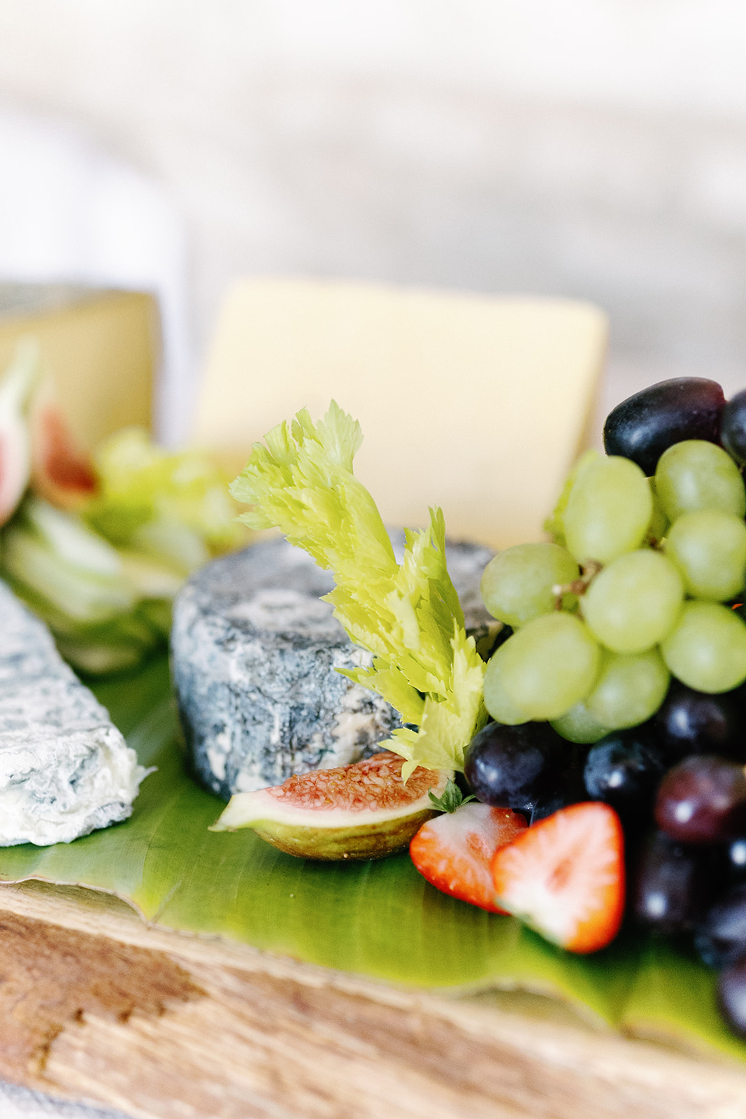 A cheese platter with assorted cheeses, green and red grapes, strawberries, celery, and figs arranged on a wooden board with a green leaf underneath.