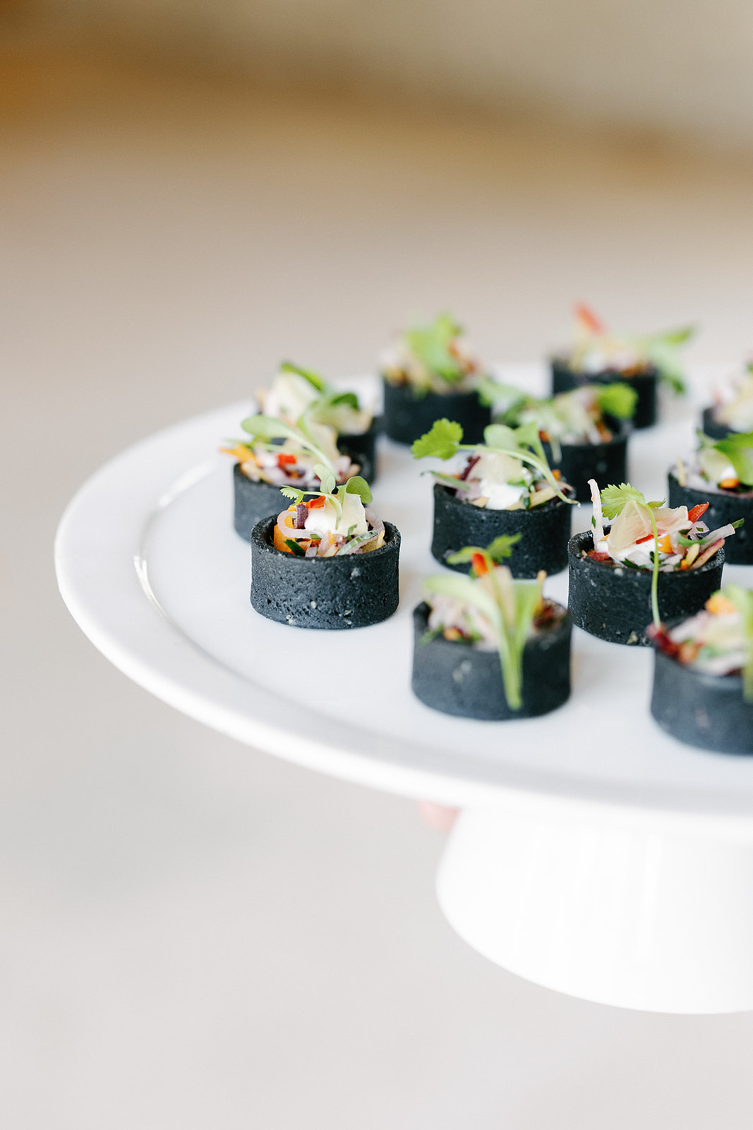 A white platter holds several black canapés topped with microgreens and garnishes, arranged in neat rows.